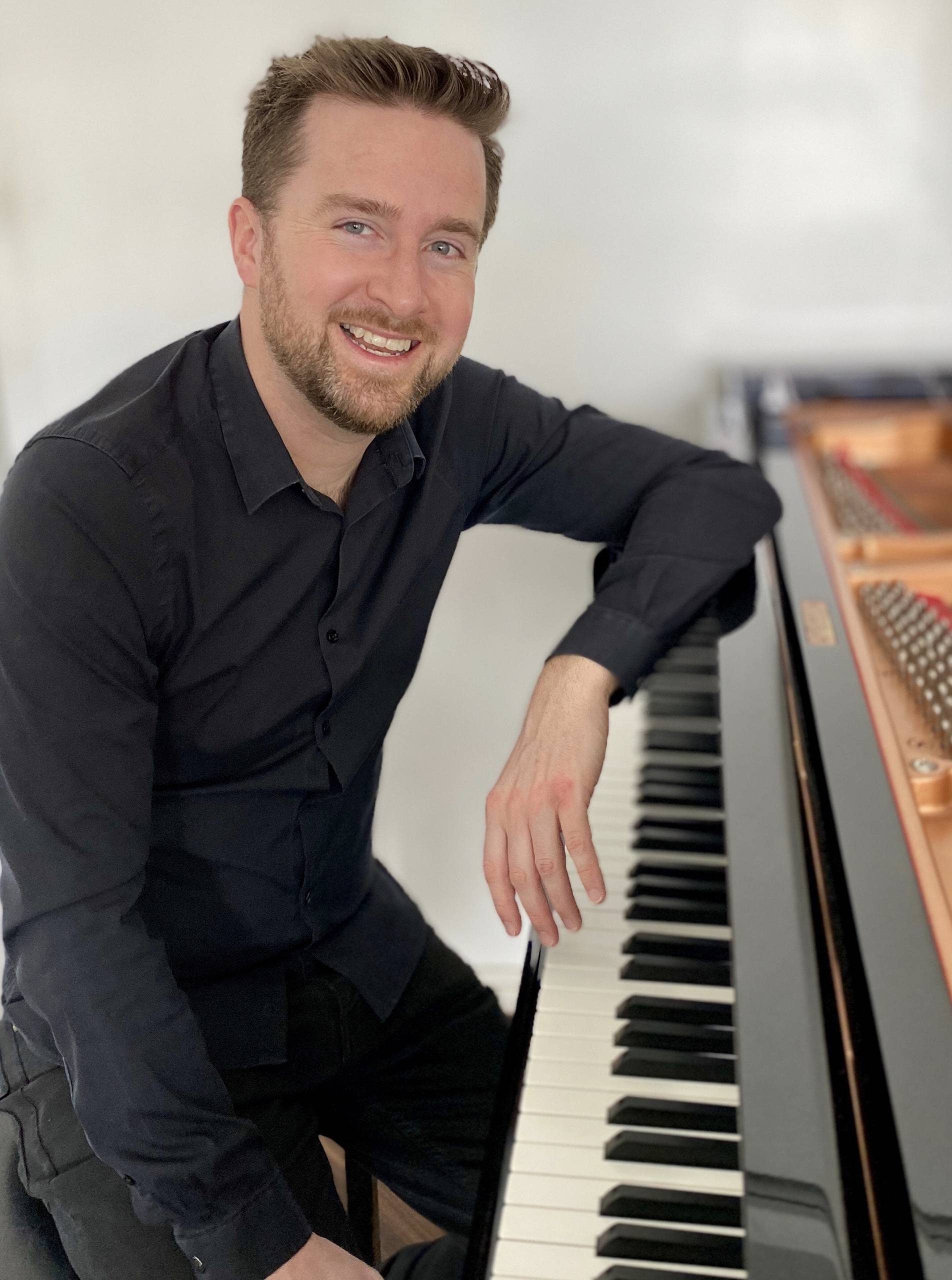 Man smiles at camera as he leans on piano.